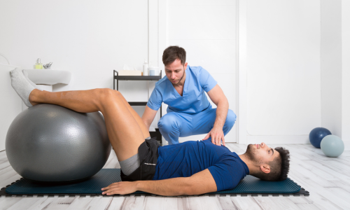 Kinésithérapeute guidant un patient allongé sur le dos, jambes posées sur un ballon de Pilates, bras le long du corps